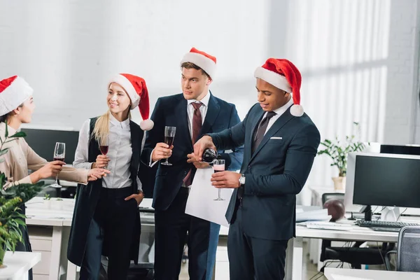 Sorrindo jovens colegas multiétnicos em chapéus de santa celebrando o Natal no escritório — Fotografia de Stock