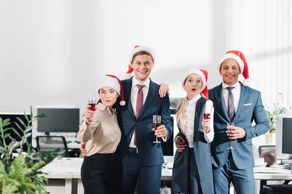 Счастливые молодые многонациональные коллеги, держащие бокалы вина и улыбающиеся перед камерой во время празднования нового года в офисе — стоковое фото
