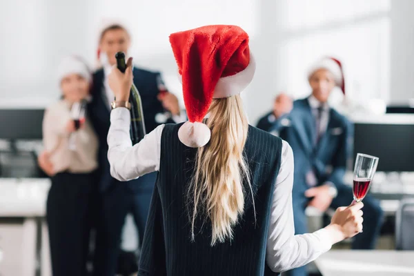 Vue arrière de la femme d'affaires dans le chapeau santa tenant verre et bouteille de champagne tout en célébrant Noël avec des collègues — Photo de stock