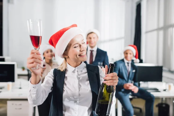 Jóvenes empresarios felices en sombreros de santa bebiendo vino y celebrando la Navidad en la oficina - foto de stock