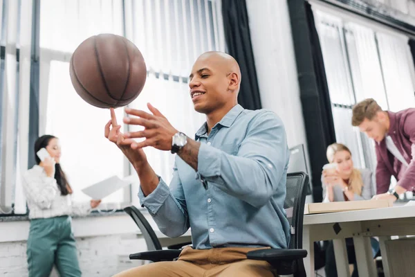 Vista basso angolo di sorridente giovane uomo africano americano che gioca con pallone da basket in ufficio — Foto stock