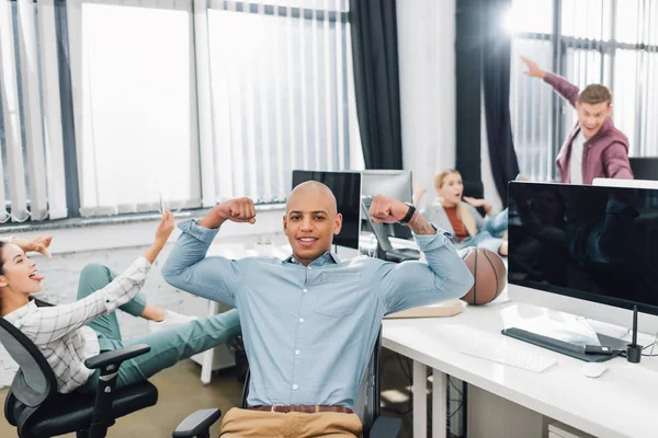 Счастливый молодой африканский американец, показывающий бицепсы и улыбающийся в камеру, в то время как коллеги развлекаются в офисе — стоковое фото
