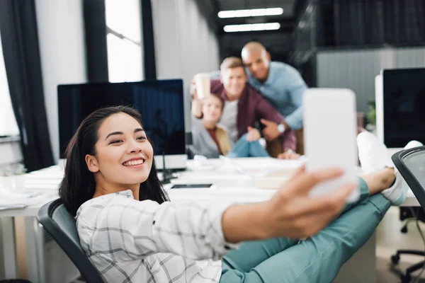 Sonriente joven empresaria tomando selfie con smartphone en oficina de espacio abierto - foto de stock