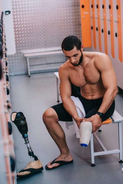 Musculoso joven deportista con pierna artificial sentado en el banco en el vestuario de la piscina - foto de stock