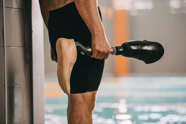 Cropped shot of swimmer standing at poolside of indoor swimming pool and holding his artificial leg — Stock Photo