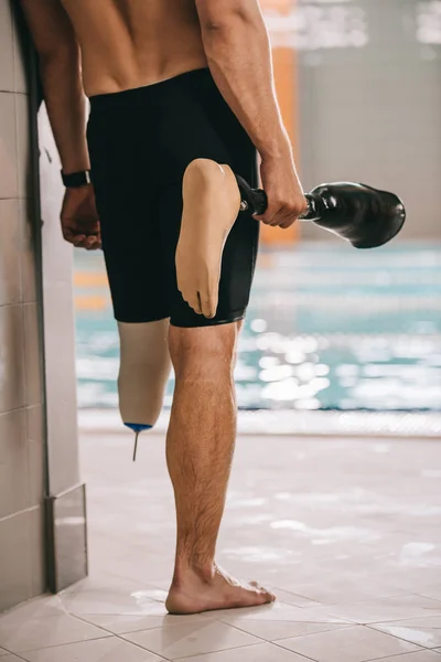 Tiro recortado de desportista em pé à beira da piscina interior e segurando sua perna artificial — Fotografia de Stock
