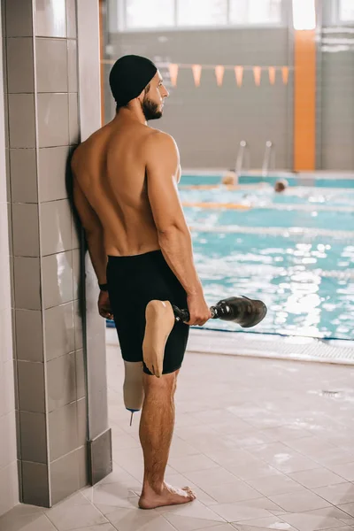 Rear view of young swimmer standing at poolside of indoor swimming pool and holding his artificial leg — Stock Photo