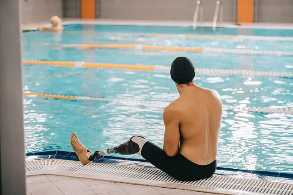 Vista posteriore del giovane nuotatore con gamba artificiale seduto a bordo piscina della piscina coperta — Foto stock