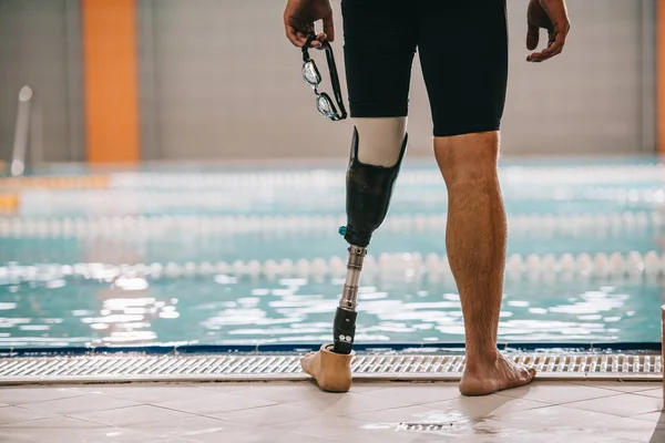 Tiro recortado de nadador com perna artificial em pé em frente à piscina interior e segurando óculos de natação — Fotografia de Stock