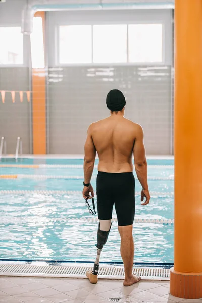 Back view of young sportsman with artificial leg looking at indoor swimming pool — Stock Photo