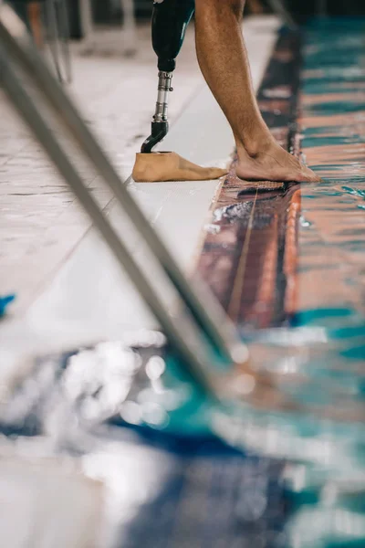Cropped shot of swimmer with artificial leg standing on poolside at indoor swimming pool and checking water temperature — Stock Photo