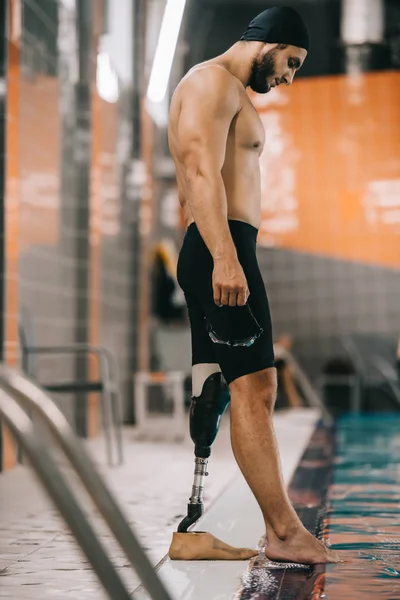 Apuesto nadador joven con pierna artificial de pie junto a la piscina en la piscina cubierta y comprobar la temperatura del agua - foto de stock