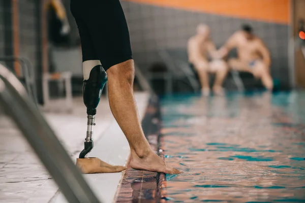 Cropped shot of sportsman with artificial leg standing on poolside at indoor swimming pool and checking water temperature — Stock Photo