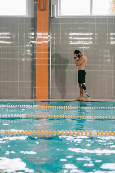 Handsome young sportsman with artificial leg walking by poolside at indoor swimming pool — Stock Photo