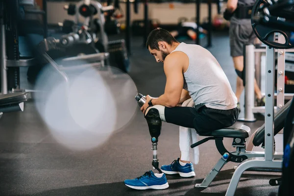 Deportista joven sobrecargado de trabajo con pierna artificial sentado en el gimnasio - foto de stock