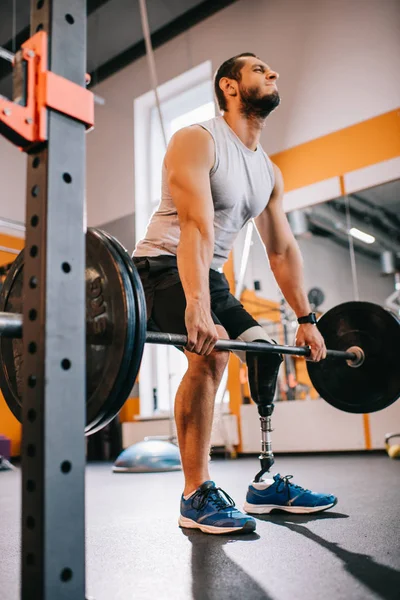 Vista inferior del joven deportista con pierna artificial haciendo ejercicio con barra en el gimnasio - foto de stock