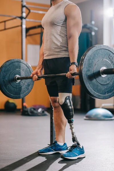 Обрезанный кадр мускулистого спортсмена с искусственной ногой, тренирующегося с штангой в спортзале — стоковое фото