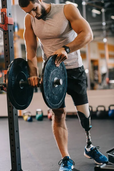 Apuesto joven deportista con pierna artificial preparándose para el entrenamiento con barra de pesas en el gimnasio - foto de stock