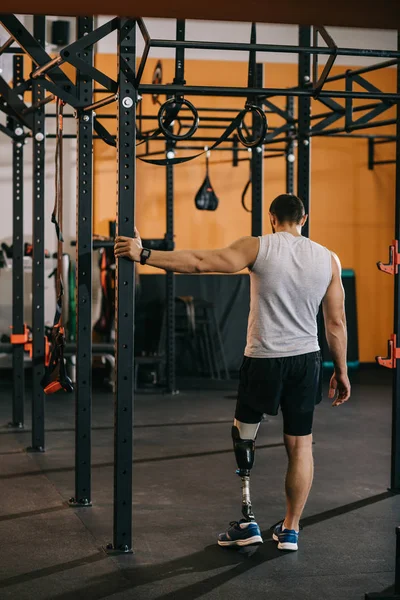 Vista posteriore del giovane sportivo con gamba artificiale in piedi vicino alla scala di ginnastica in palestra — Foto stock