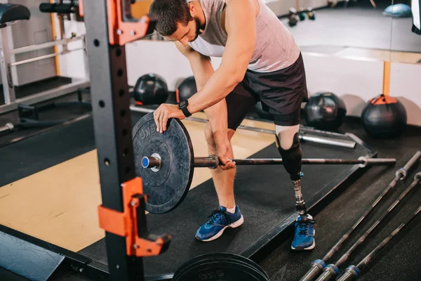 Handsome young sportsman with artificial leg putting on weight plate on bar at gym before workout — Stock Photo