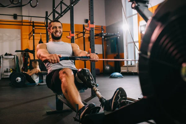 Bello giovane sportivo con gamba artificiale che lavora fuori con vogatore in palestra — Foto stock