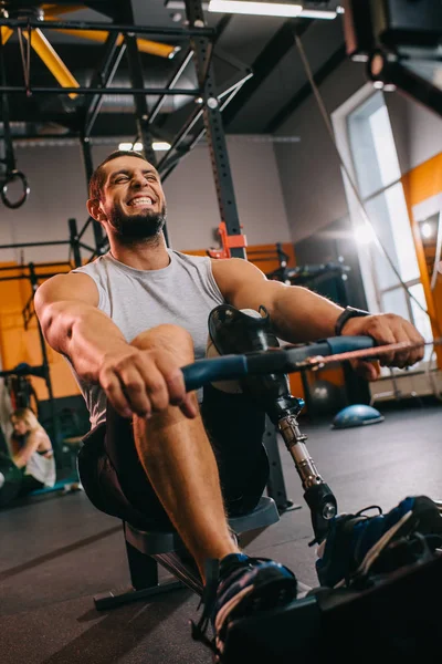 Hardworking young sportsman with artificial leg working out with rowing machine at gym — Stock Photo