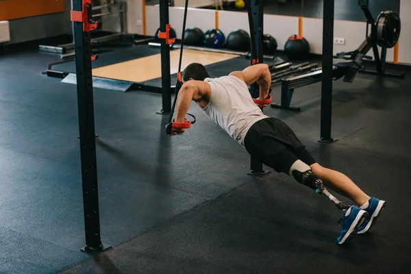 Красивый молодой спортсмен с искусственной ногой делает отжимания с висящими кольцами лестницы гимнастики в тренажерном зале — стоковое фото