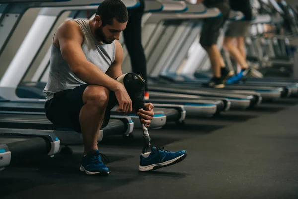 Beau jeune sportif avec jambe artificielle assis sur tapis roulant à la salle de gym — Photo de stock