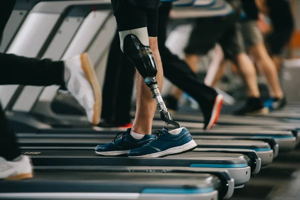 Tiro recortado de deportista con pierna artificial corriendo en la cinta de correr en el gimnasio con otras personas - foto de stock