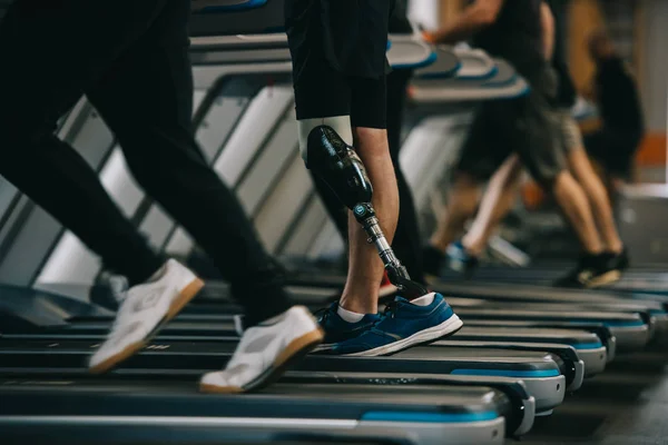 Colpo ritagliato di sportivo con gamba artificiale che corre sul tapis roulant in palestra con altre persone — Foto stock