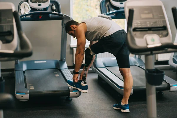 Атлетичный молодой спортсмен с искусственной ногой, зашнуривающий обувь в спортзале — стоковое фото