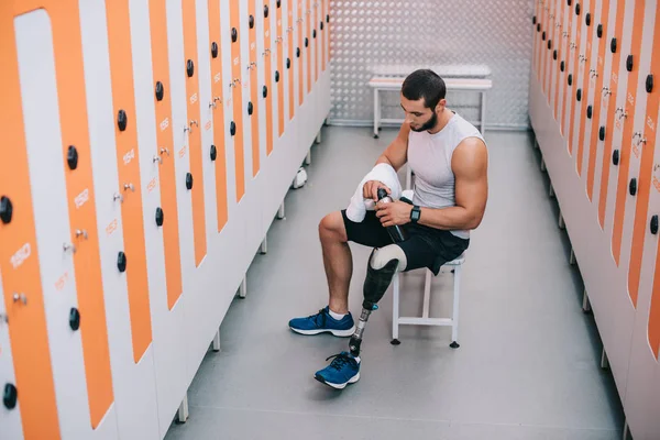 Handsome young sportsman with artificial leg sitting on bench at gym changing room — Stock Photo