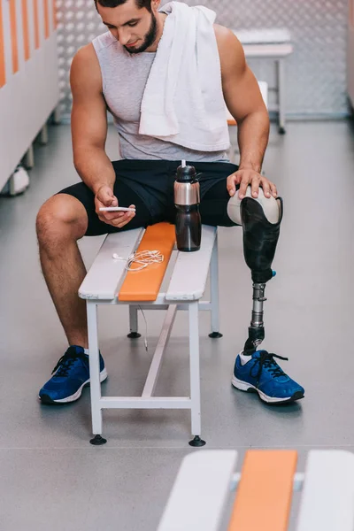 Attractive young sportsman with artificial leg sitting on bench at gym changing room and using smartphone — Stock Photo