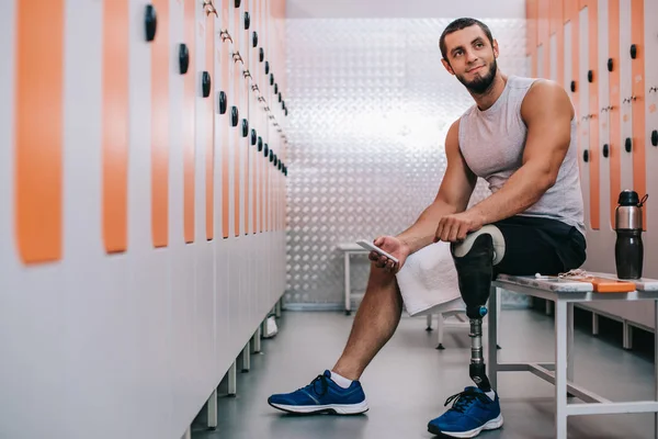 Усміхнений молодий спортсмен зі штучною ногою сидить на лавці в роздягальні спортзалу і використовує смартфон — стокове фото