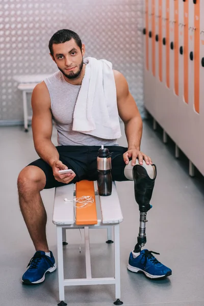 Beau jeune sportif avec jambe artificielle assis sur le banc à la salle de gym vestiaire et en utilisant smartphone — Photo de stock