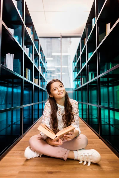 Belle écolière souriante tenant le livre et regardant loin tout en étant assis dans le sol dans la bibliothèque — Photo de stock