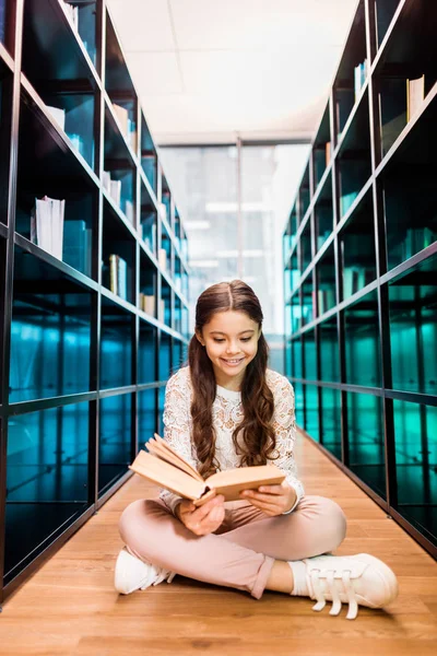 Hermosa colegiala sonriente sentada en el suelo y leyendo libro en la biblioteca - foto de stock