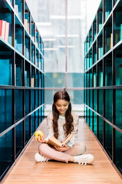 Adorable happy girl holding apple and reading book in library — Stock Photo