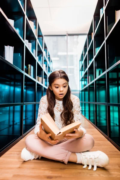 Adorable impactado colegiala sentado en el suelo y lectura de libro en la biblioteca - foto de stock