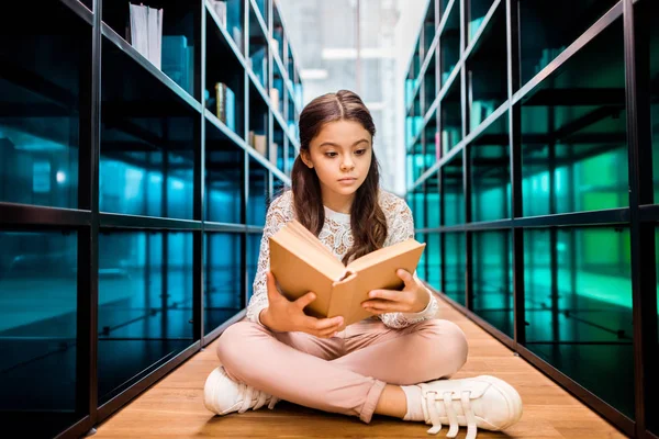 Adorable écolière concentrée assise sur le sol et le livre de lecture dans la bibliothèque — Photo de stock
