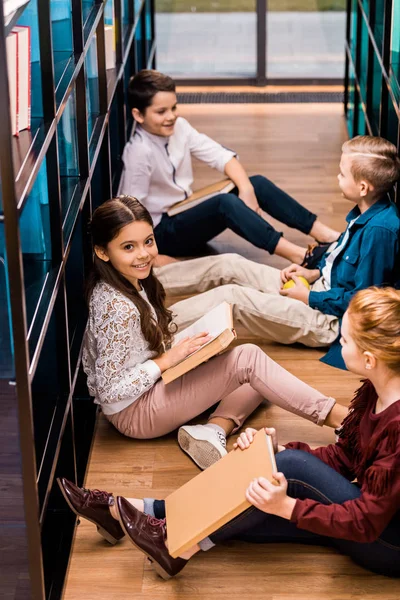 Высокий угол обзора четырех одноклассников, сидящих на полу и читающих книги в библиотеке — стоковое фото