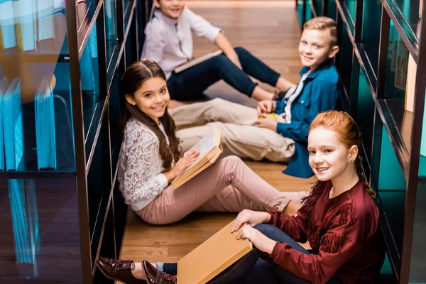 Высокий угол обзора одноклассников с книгами, сидящими на полу и улыбающимися перед камерой в библиотеке — стоковое фото