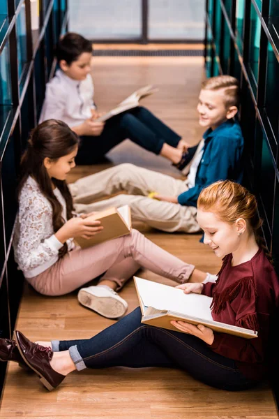 Vista de ángulo alto de cuatro escolares sentados en el suelo y leyendo libros en la biblioteca — Stock Photo