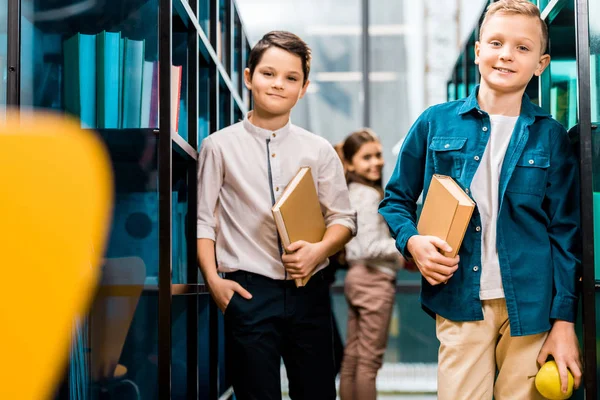 Adorable schoolchildren holding books and smiling at camera in library — Stock Photo