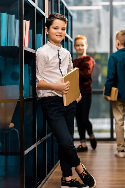 Scolaro che tiene libro e sorride alla macchina fotografica mentre visita la biblioteca con i compagni di classe — Foto stock