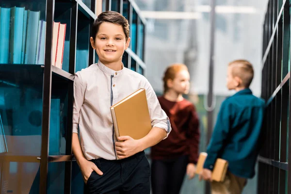 Entzückender Junge hält Buch in der Hand und lächelt in die Kamera, während er mit Klassenkameraden die Bibliothek besucht — Stockfoto