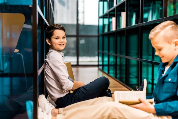 Seitenansicht von entzückend lächelnden Schuljungen, die Bücher lesen, während sie auf dem Boden in der Bibliothek sitzen — Stockfoto