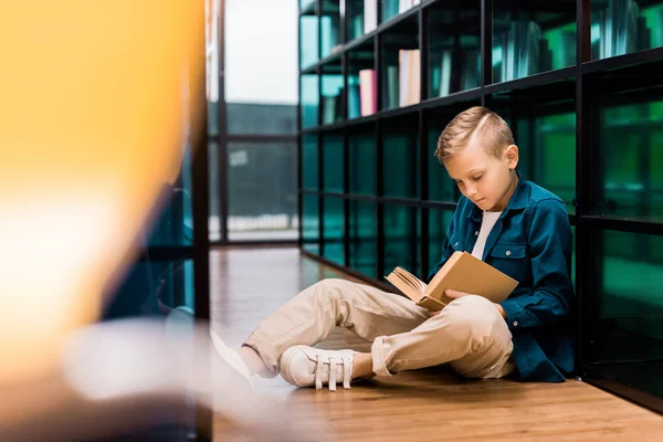 Fofo focado menino leitura livro e sentado no chão na biblioteca — Fotografia de Stock