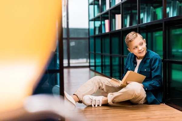 Netter lächelnder Junge liest Buch und schaut weg, während er auf dem Boden in der Bibliothek sitzt — Stockfoto