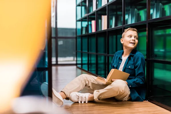 Sonriente colegial sosteniendo el libro y mirando hacia otro lado mientras está sentado en el suelo en la biblioteca - foto de stock